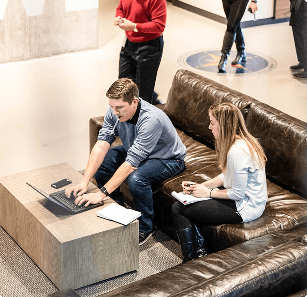 Overhead view of two colleagues working together on a leather couch, one typing on a laptop and one taking notes