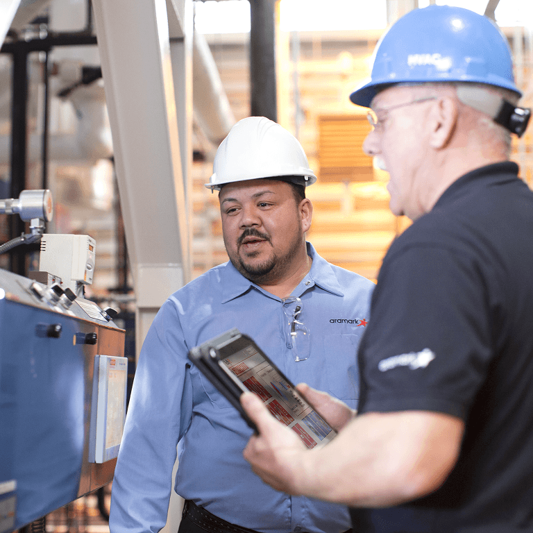 Two Aramark engineers in hard hats reviewing equipment data on a tablet in a mechanical room