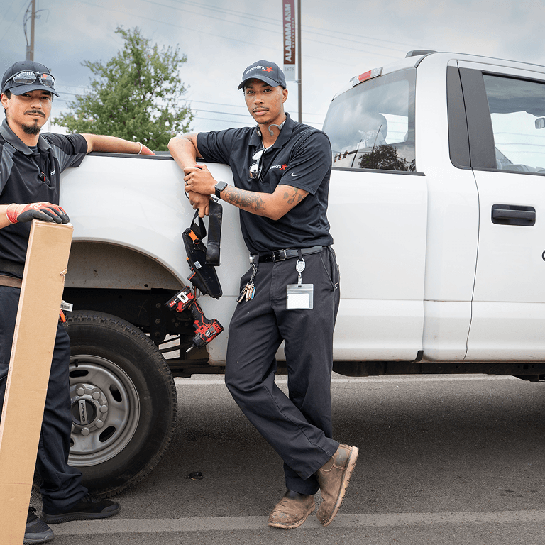 Two Aramark facilities technicians with tool belts leaning against a white pickup truck