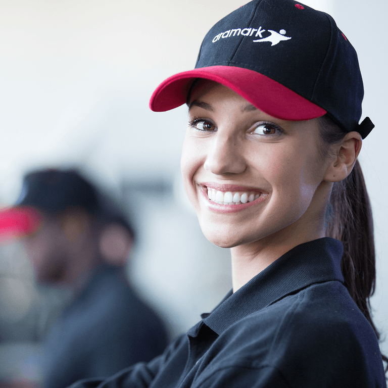 Smiling Aramark staff member wearing a navy and red branded cap, looking over her shoulder at the camera