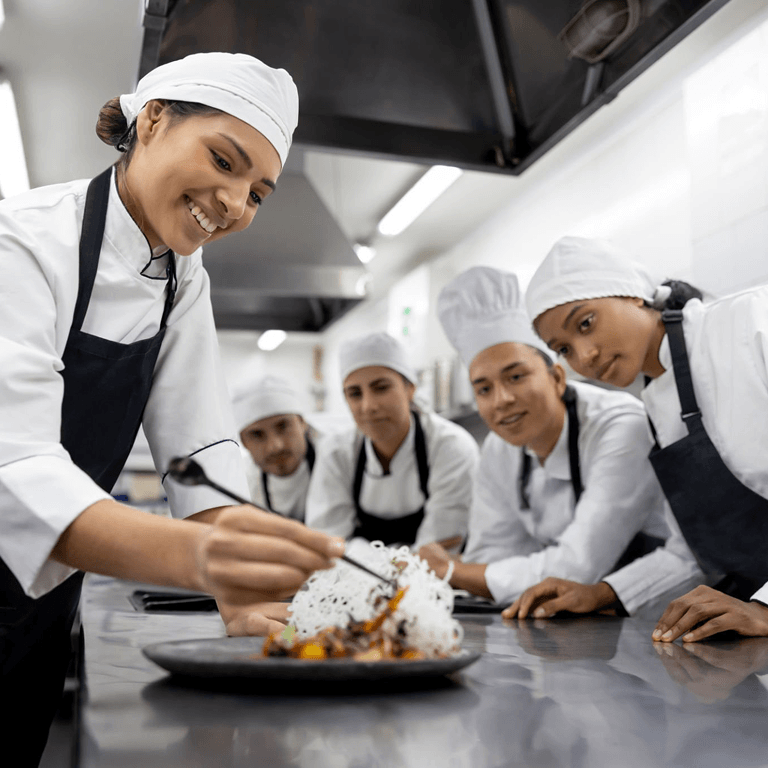 Chef instructor plating a dish while culinary students in chef whites look on in a training kitchen