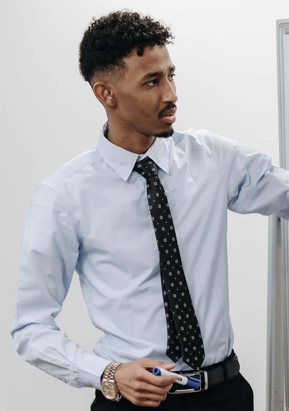 Man in a light blue dress shirt and patterned tie standing beside a whiteboard, holding a marker