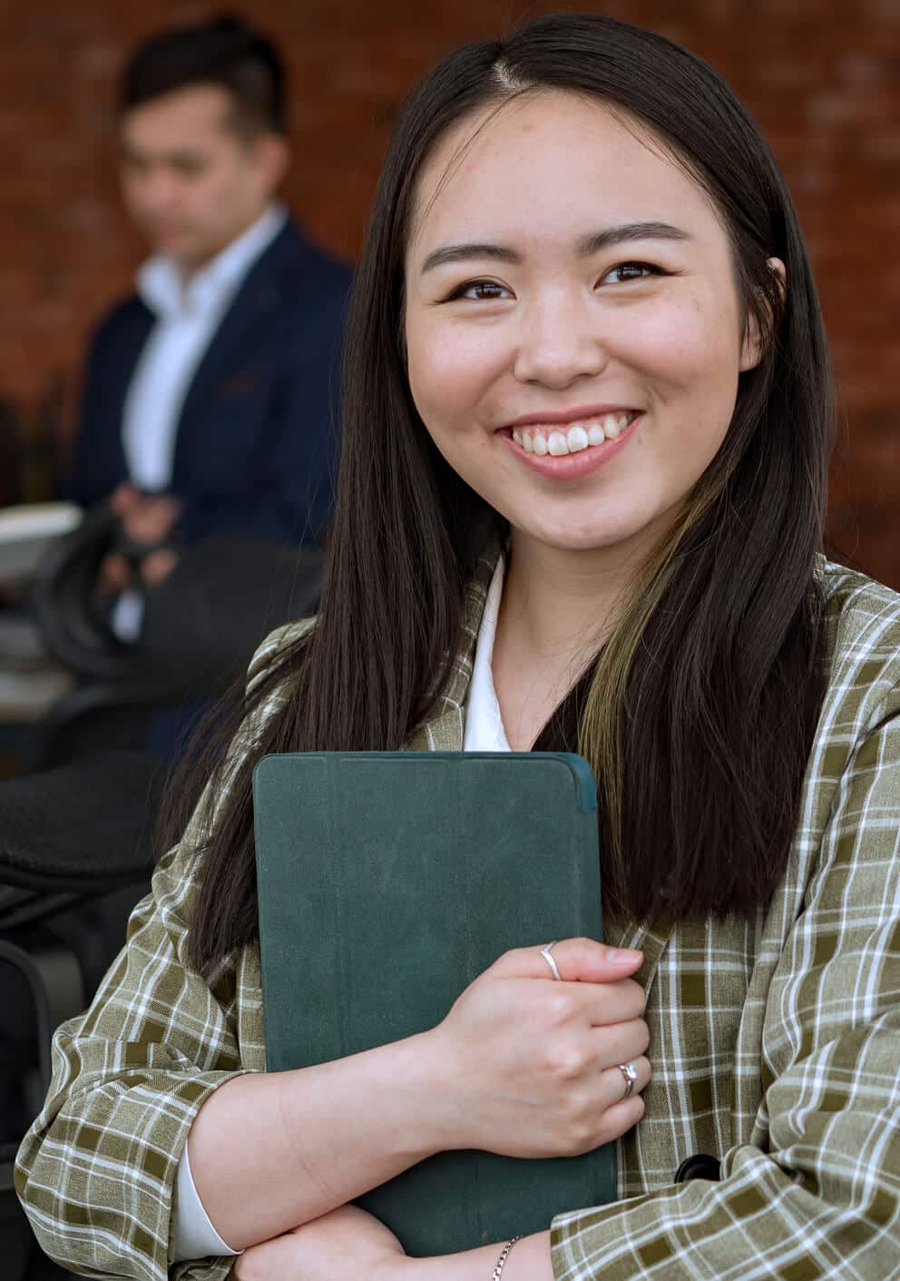 Smiling woman in a plaid jacket holding a tablet, seated outdoors with a colleague in the background