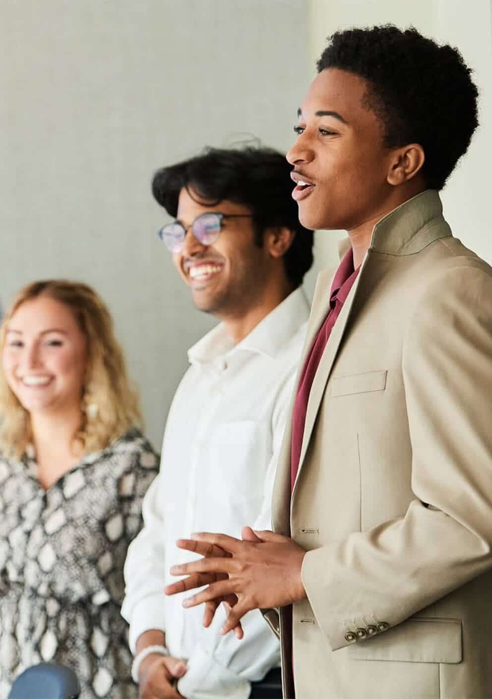 Young professional in a tan blazer speaking while two smiling colleagues stand beside him