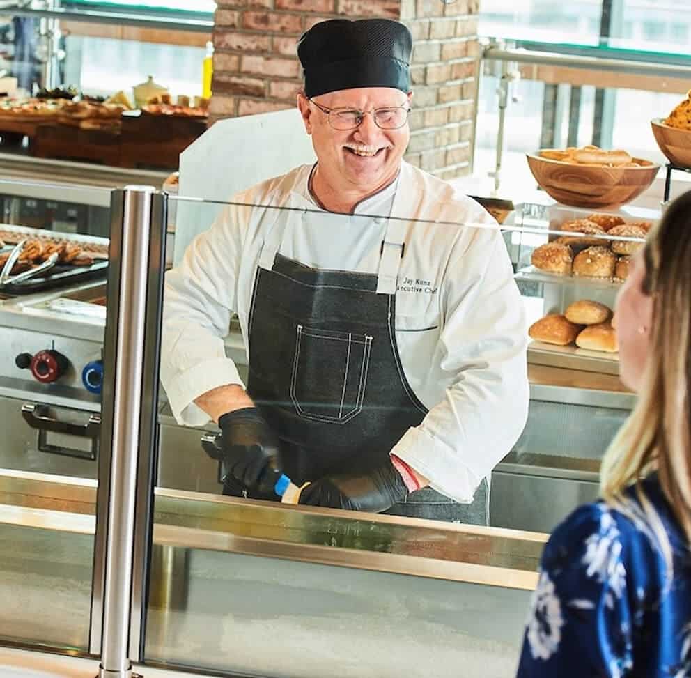 Smiling executive chef in a black cap and apron greeting a customer at a bakery counter filled with fresh bread