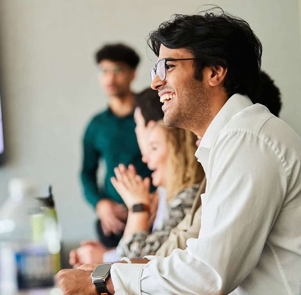 Man in glasses smiling in a meeting while colleagues in the background react and applaud