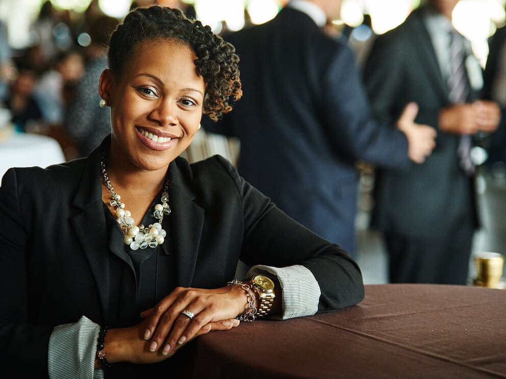 Smiling woman in a black blazer seated at a table during an indoor event with colleagues in the background
