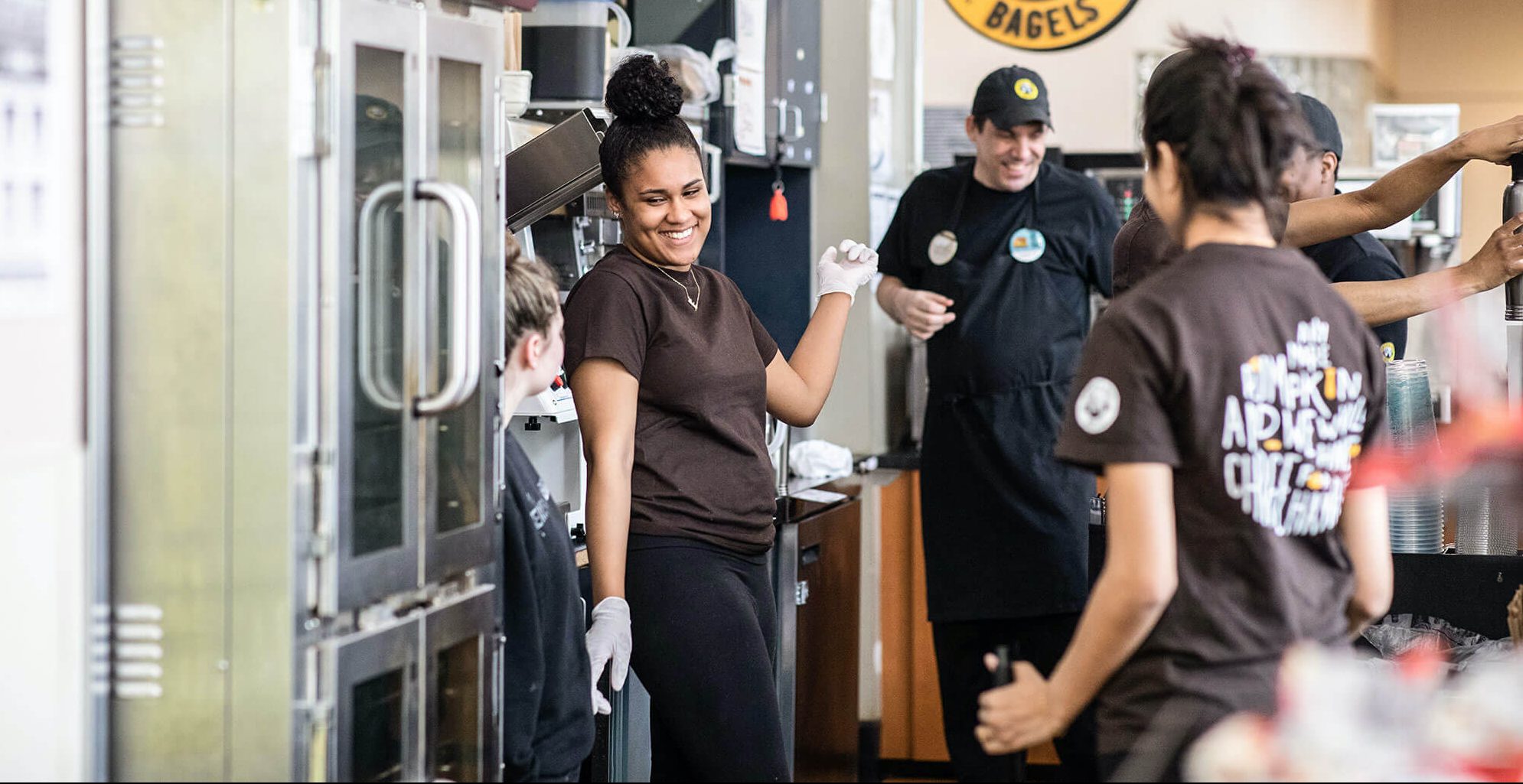 Smiling staff members in brown shirts working together behind the counter of a bagel shop